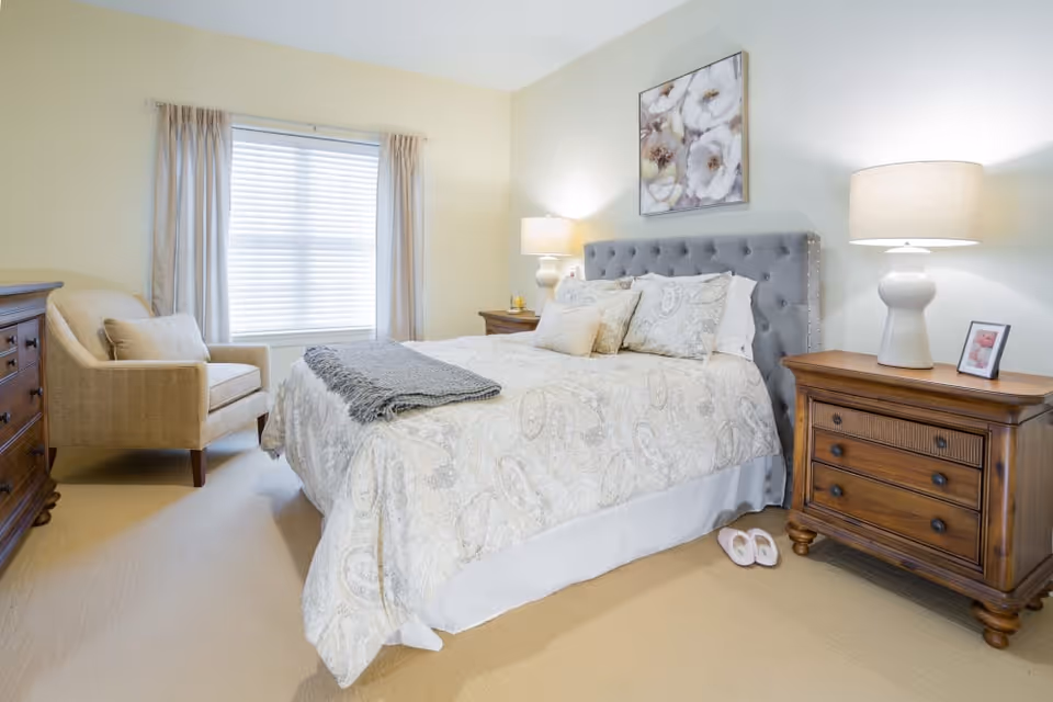 A bright and cozy bedroom with a large bed featuring a tufted gray headboard and patterned bedding. There are two wooden nightstands on either side of the bed, each with a lamp and decorative items. A beige armchair is placed near a window with closed blinds and light curtains. The room has a soft beige carpet and light-colored walls, with a floral painting hanging above the bed.