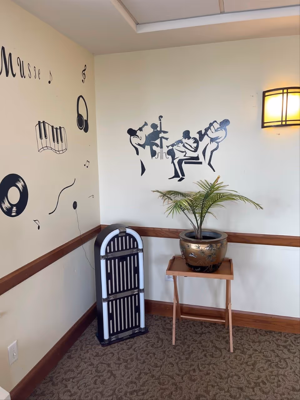 Corner of a room with musical-themed wall decals including a piano keyboard, headphones, a vinyl record, musical notes, and silhouettes of jazz musicians playing instruments. There is a vintage-style jukebox on the floor and a potted plant on a wooden stand. A wall-mounted light fixture is on the right side.