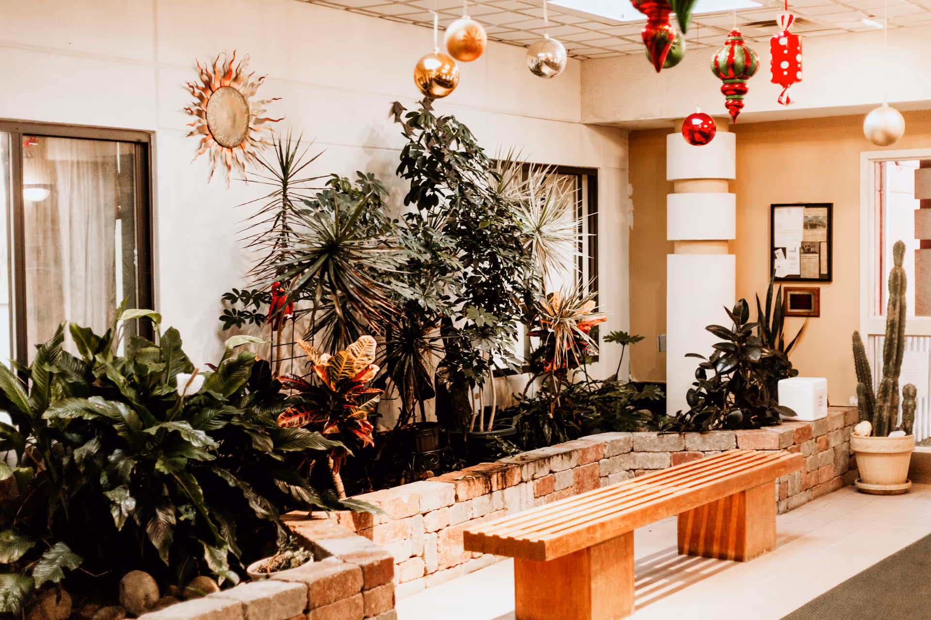 Indoor seating area with a wooden bench in front of a raised stone planter filled with various green plants. The wall behind has windows and a decorative sun sculpture. Festive hanging ornaments are suspended from the ceiling.
