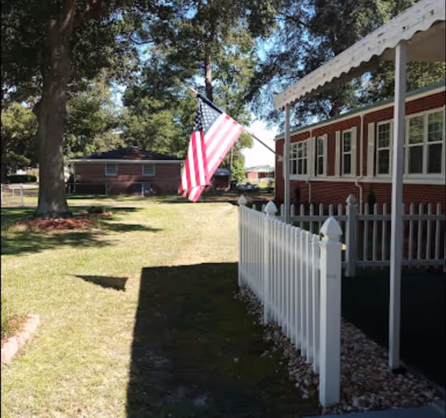 Outdoor view of a residential area with a white picket fence, a porch with a white awning, an American flag mounted on the side of a red brick building, large trees, and a grassy yard with another brick building in the background.