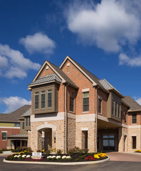 Brick-and-stone front of a senior living building with an arched entrance, landscaped flowerbeds and a sign reading "The Grand" under a blue sky.