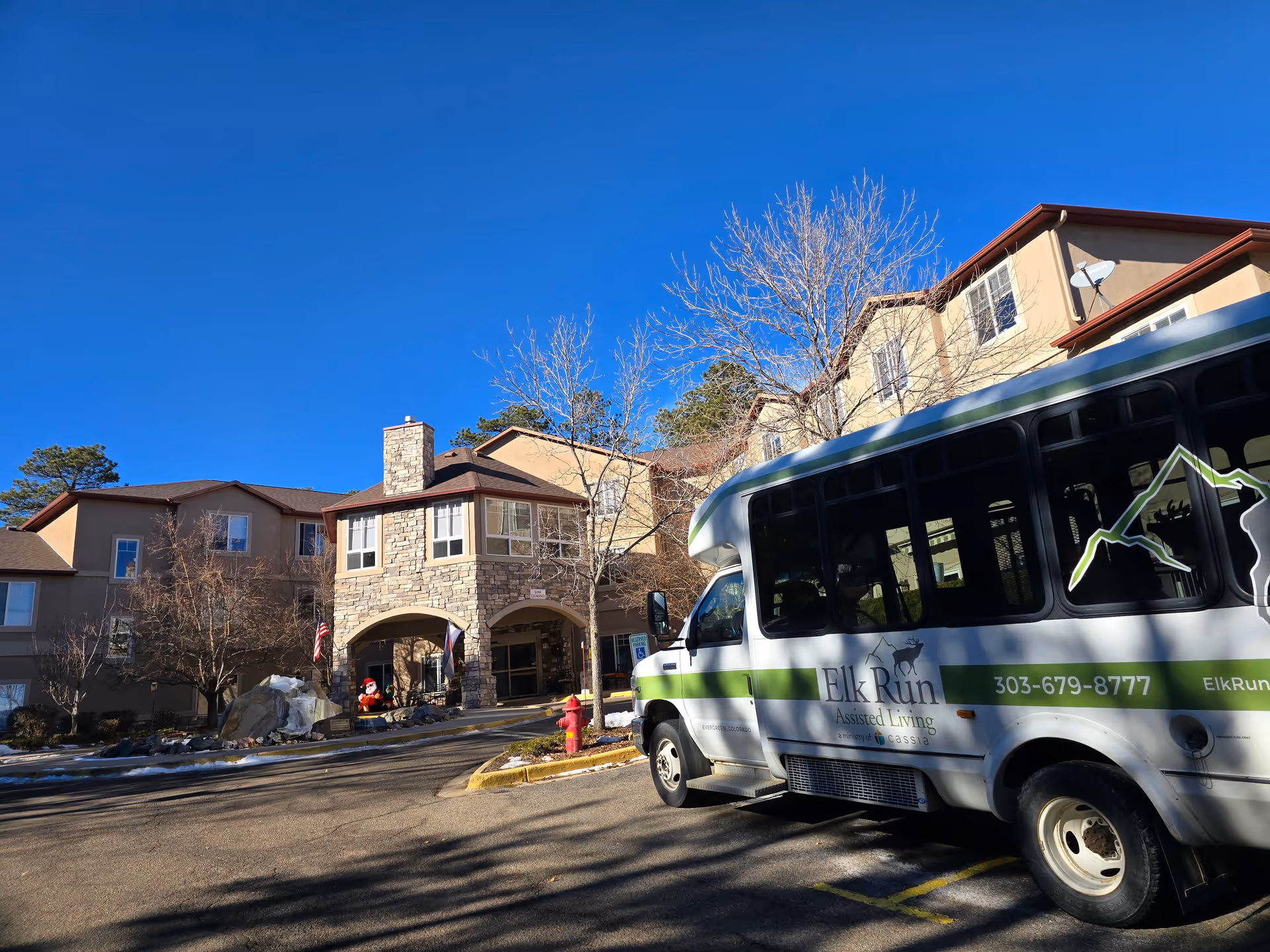 Exterior view of Elk Run Assisted Living facility on a clear sunny day with a shuttle bus parked in front. The building has a stone entrance with arches and multiple windows. There are leafless trees and a small rock garden near the entrance.