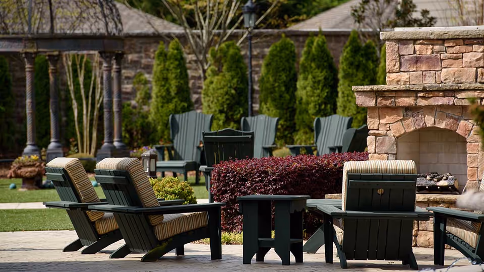 Outdoor seating area with cushioned Adirondack chairs around a stone fireplace in a landscaped garden.