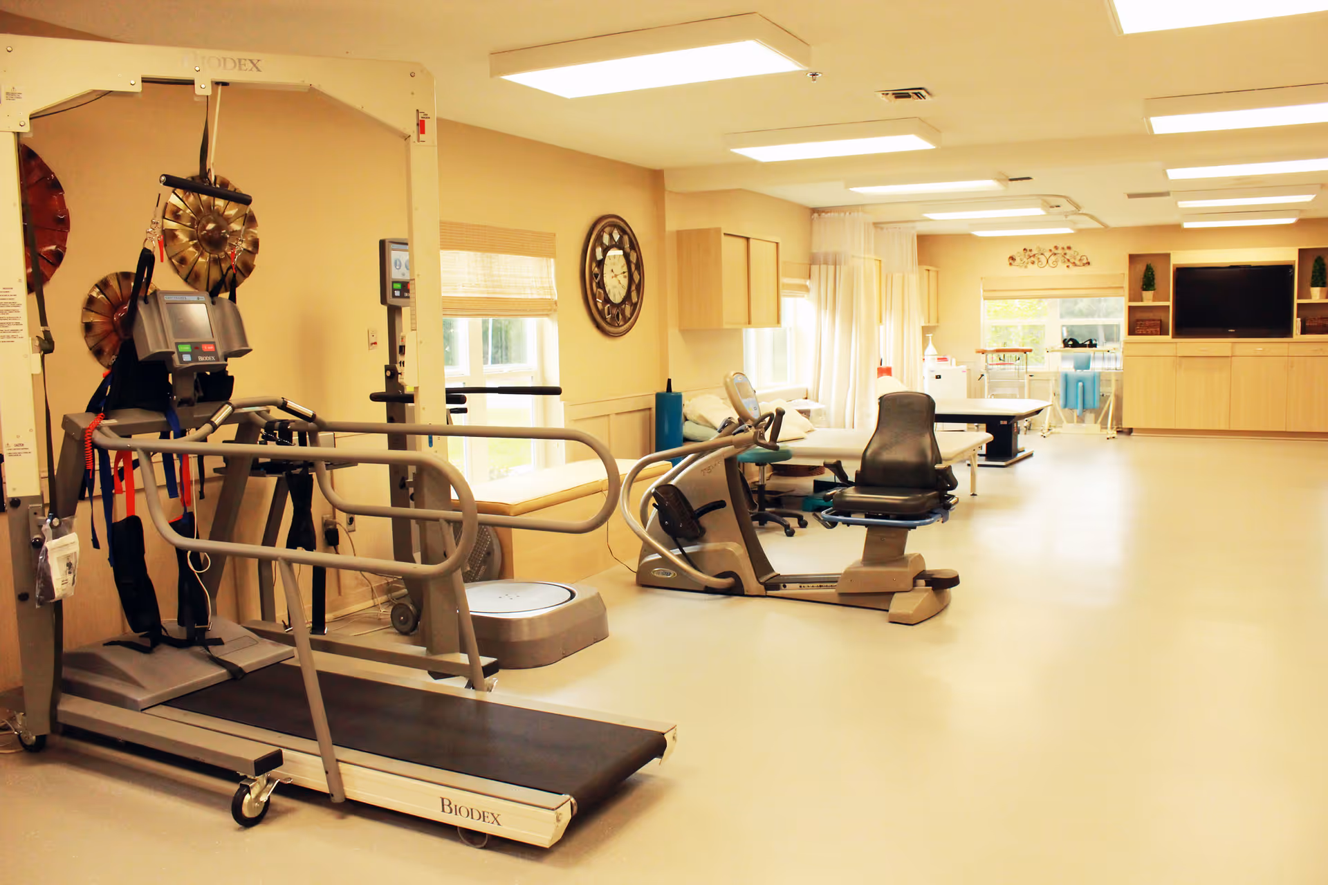 A bright rehabilitation room with exercise equipment including a treadmill, stationary bike, and therapy tables. The room has large windows, light-colored walls, and ceiling lights. There is a large clock on the wall and a television mounted on a cabinet in the background.