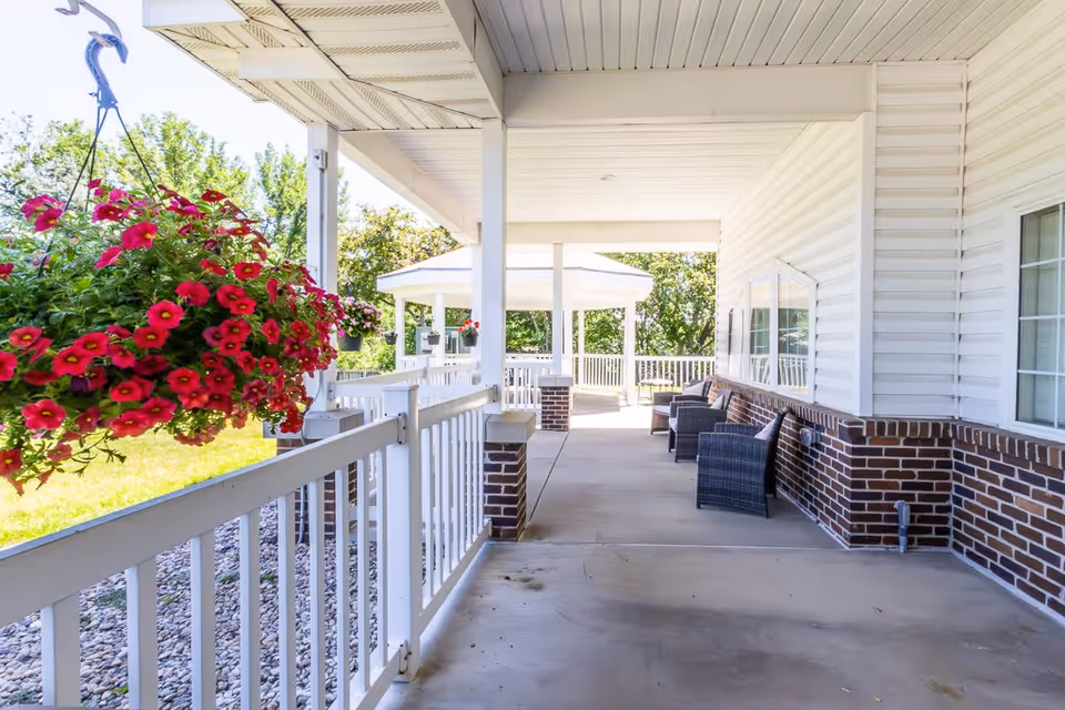 Covered outdoor patio area with white railing and brick accents, featuring hanging baskets of red flowers and several wicker chairs with cushions along the wall. Trees and greenery are visible in the background under a clear sky.
