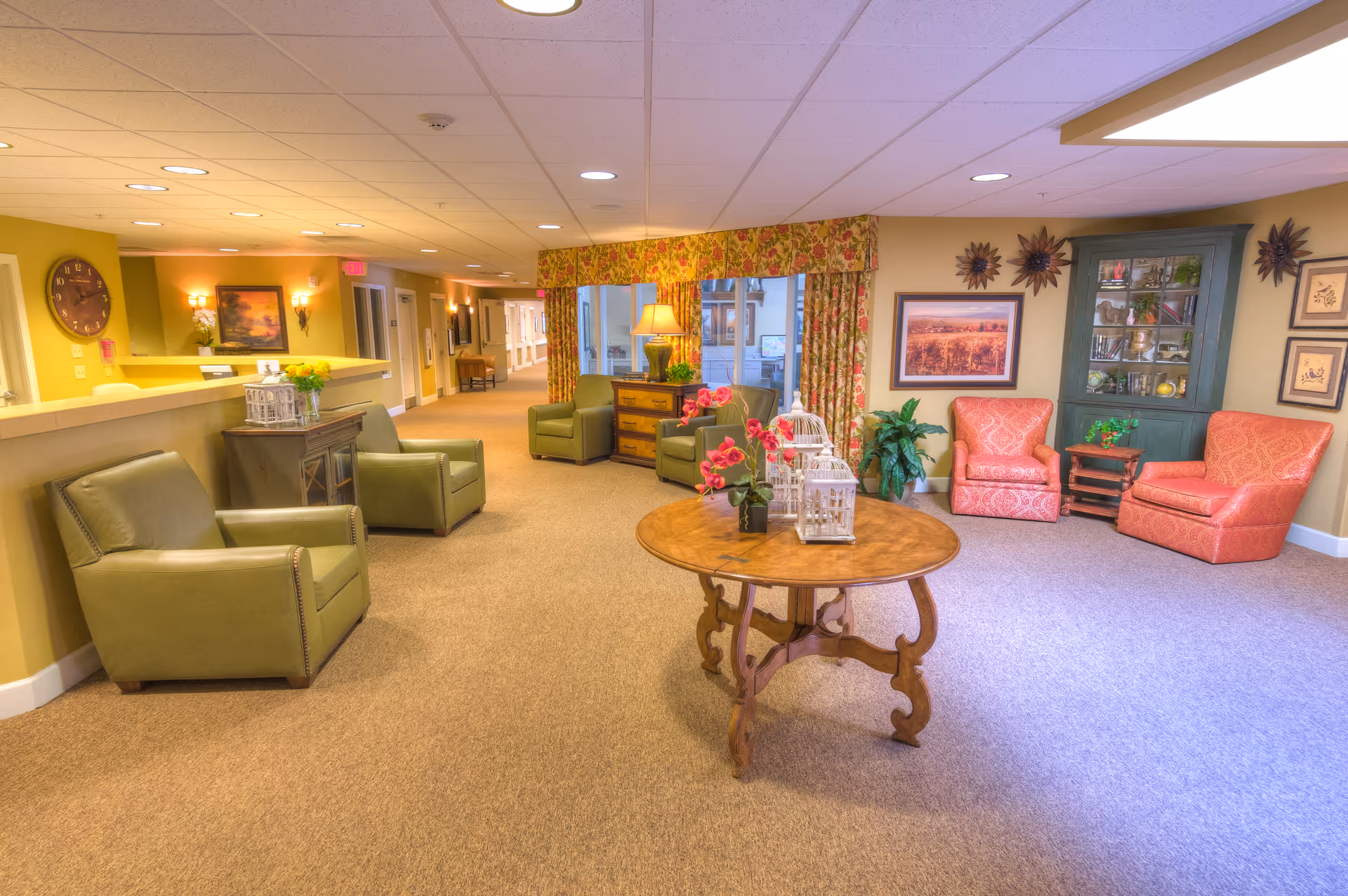 Well-lit communal sitting area with armchairs, a round wooden table and decorative furnishings in a memory care facility.