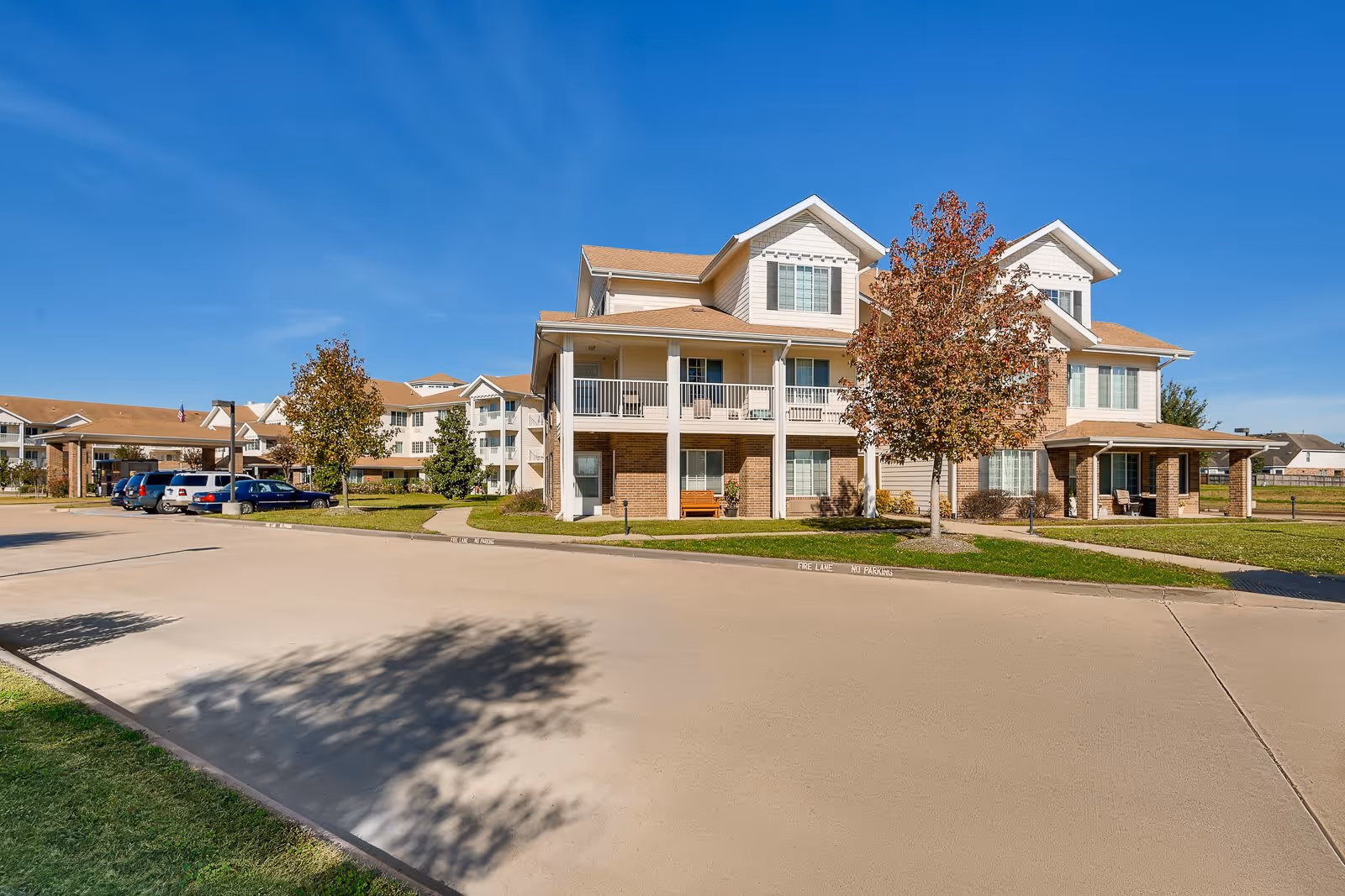Exterior view of a multi-story residential building with beige siding and brick accents, surrounded by green lawns and trees under a clear blue sky. Several parked cars are visible on the left side along a paved driveway.