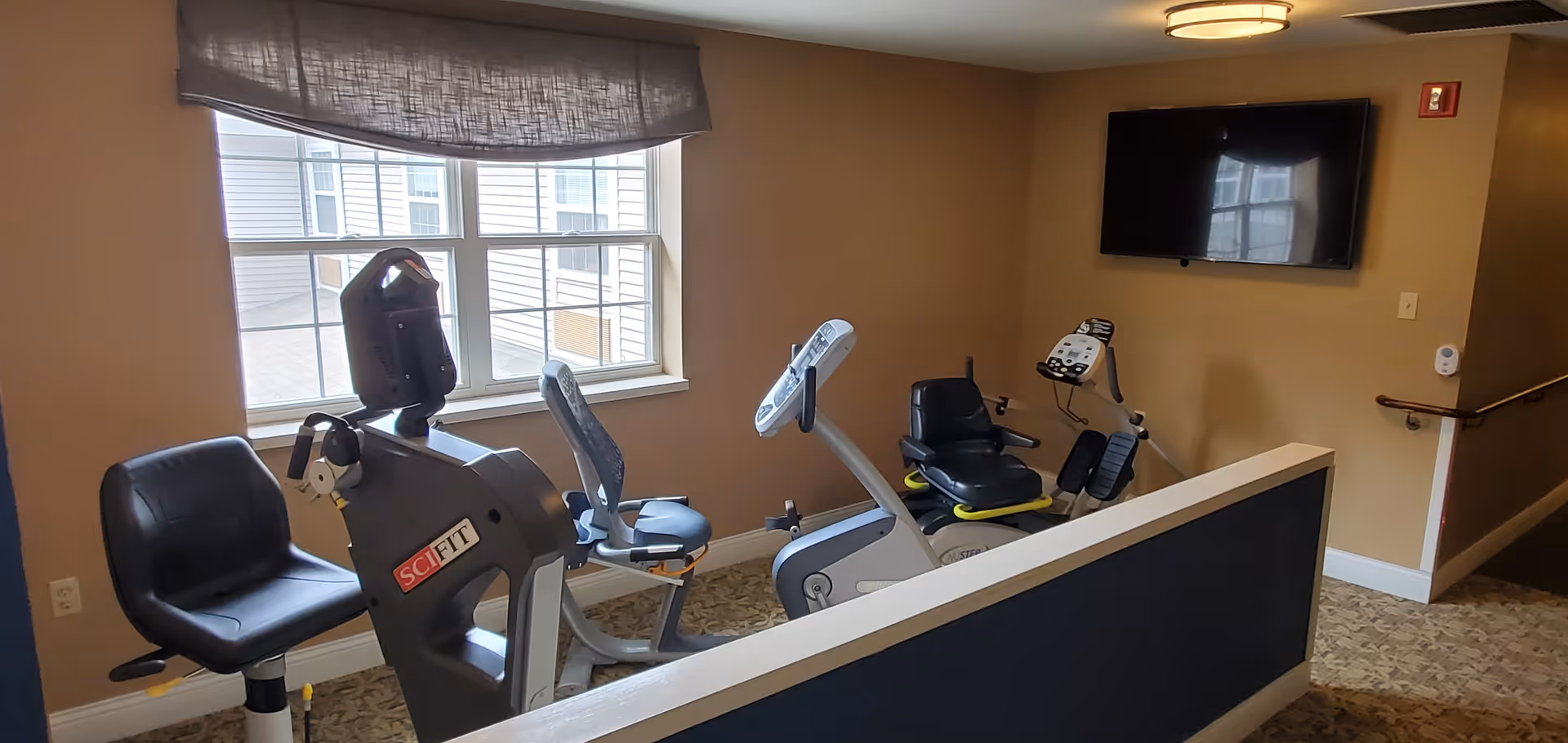 A small exercise room with two stationary exercise bikes and one recumbent bike positioned near a window. The room has beige walls, a patterned carpet, a wall-mounted TV, and a ceiling light fixture.