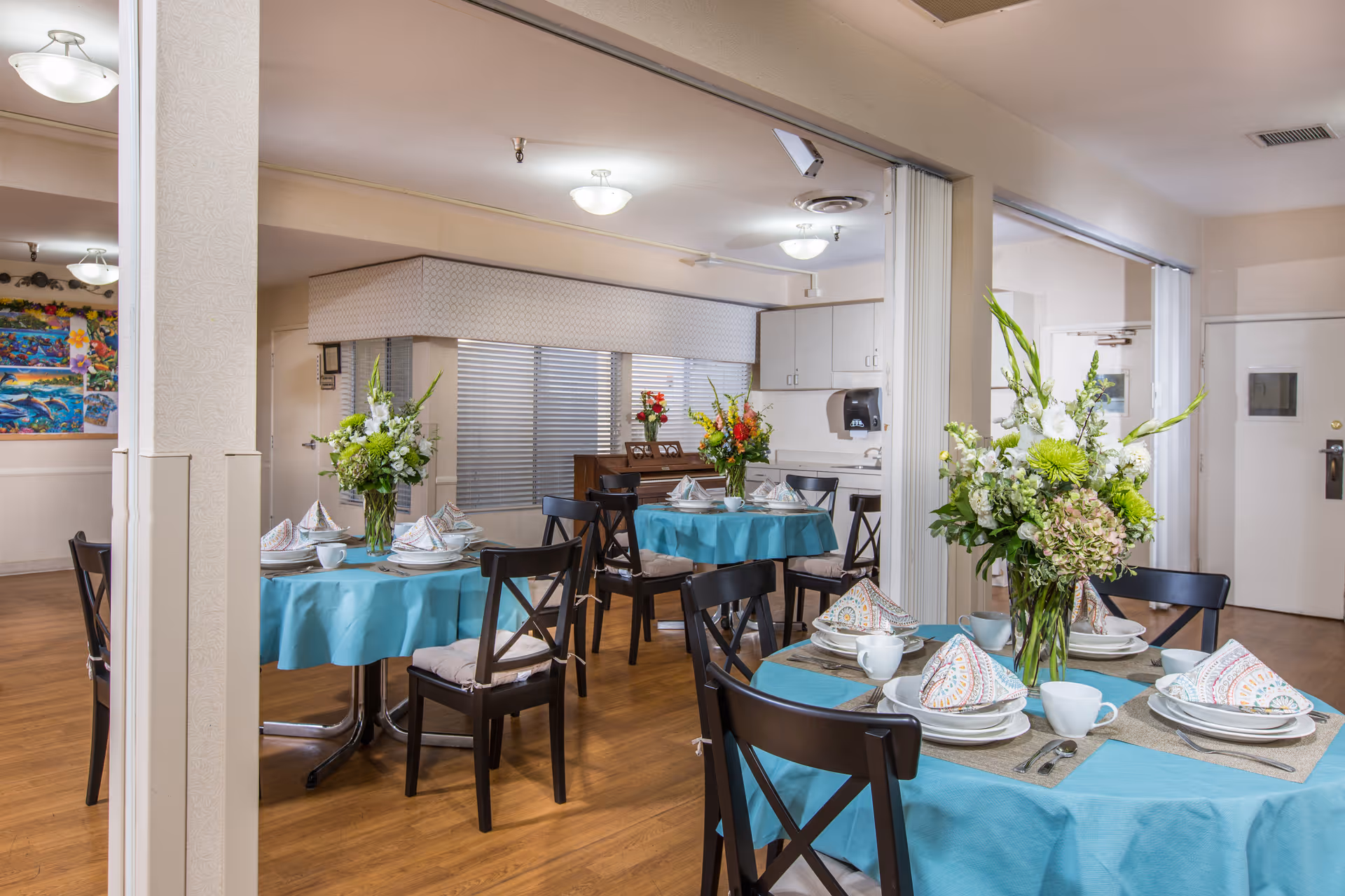 Dining area with round tables covered in blue tablecloths, each set with plates, cups, napkins, and silverware. Each table has a large floral centerpiece. The room has wooden flooring, white walls, and ceiling lights. There are black chairs around the tables and a piano in the background.