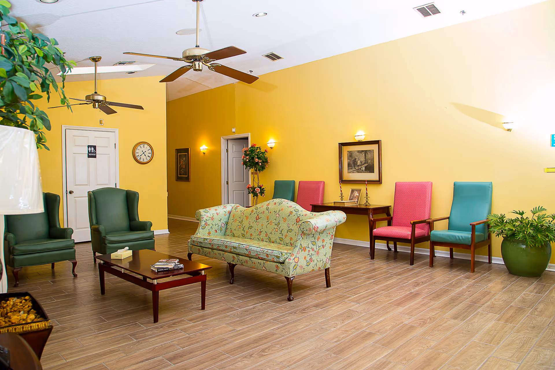 A cozy assisted living facility common area with yellow walls and wood-look tile flooring. The room features a floral patterned sofa, two green armchairs, a wooden coffee table with books and tissues, and a row of colorful chairs against the wall. Ceiling fans and wall sconces provide lighting, and there are framed pictures and plants decorating the space.