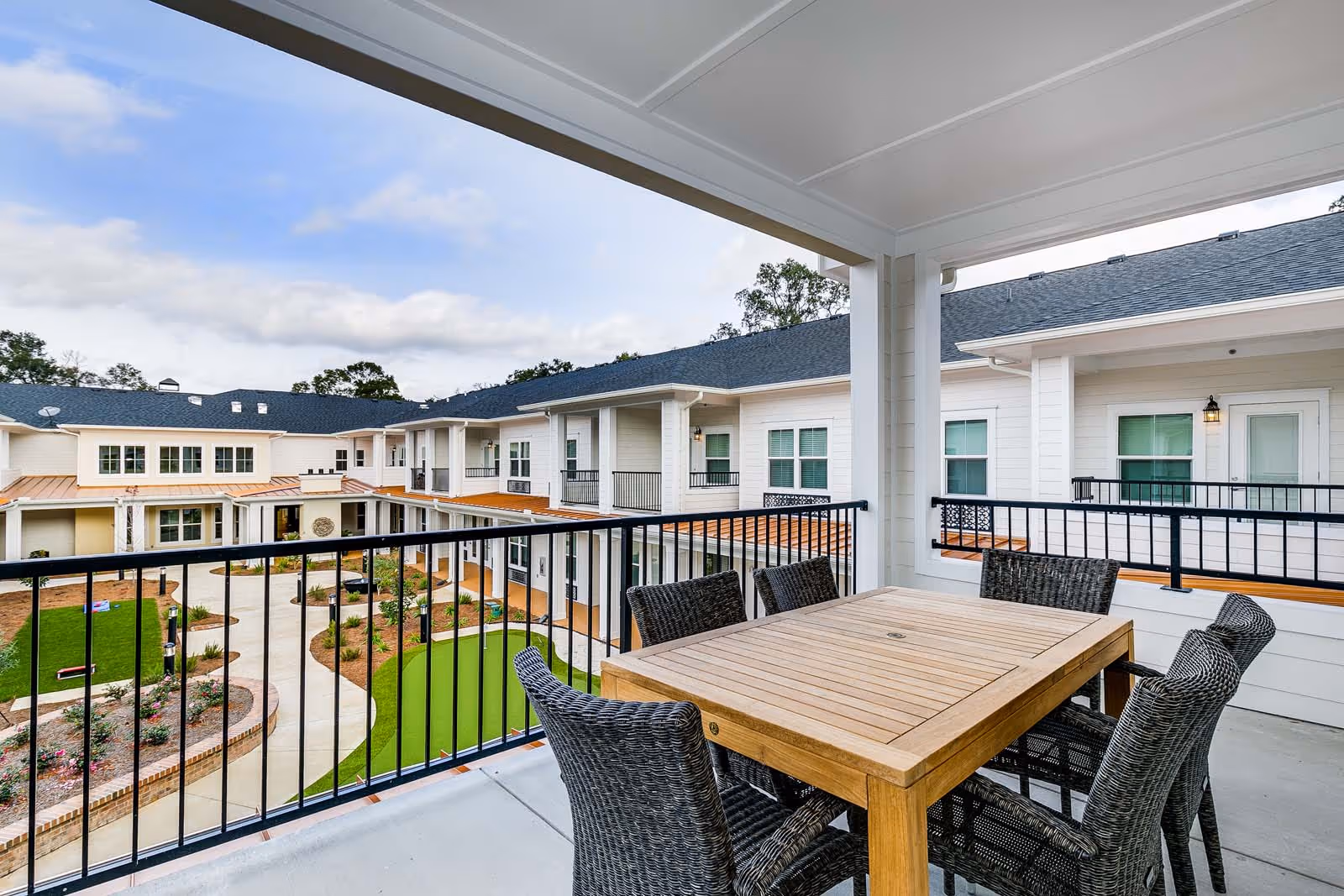 Covered balcony area with a wooden table and six wicker chairs overlooking a landscaped courtyard with pathways, greenery, and a two-story building with white siding and multiple windows.
