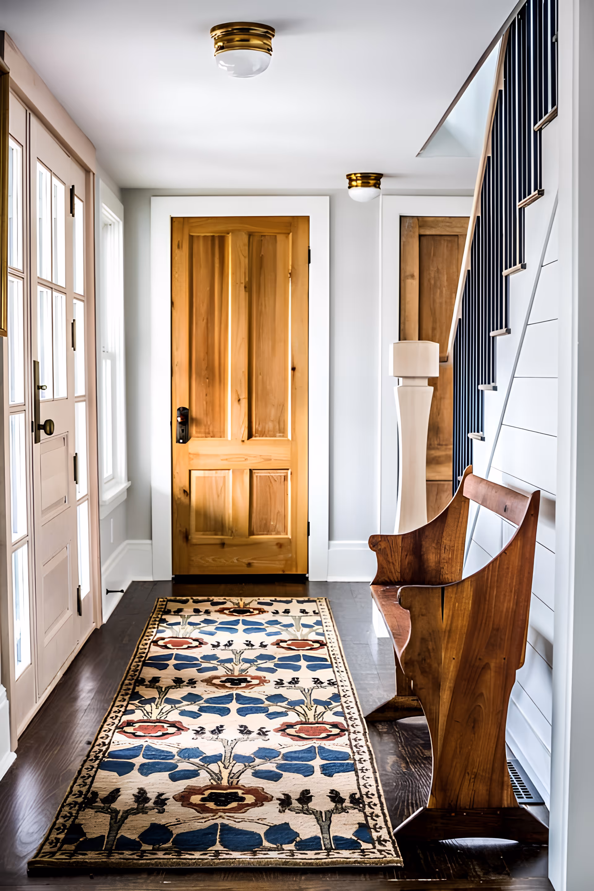 Bright entry hallway with a wooden front door, patterned runner rug, wooden bench and staircase railing.