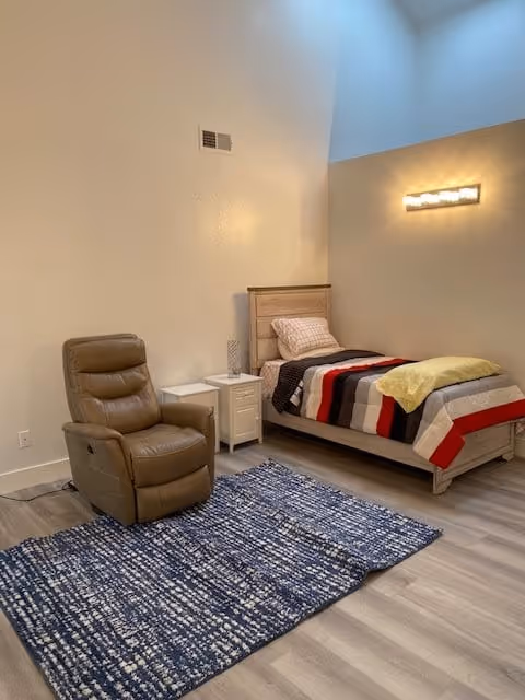 A cozy senior living bedroom featuring a single bed with a striped blanket and a yellow throw, a brown recliner chair, a white nightstand with a decorative glass, a blue patterned rug on the wooden floor, and a modern wall light fixture illuminating the room.