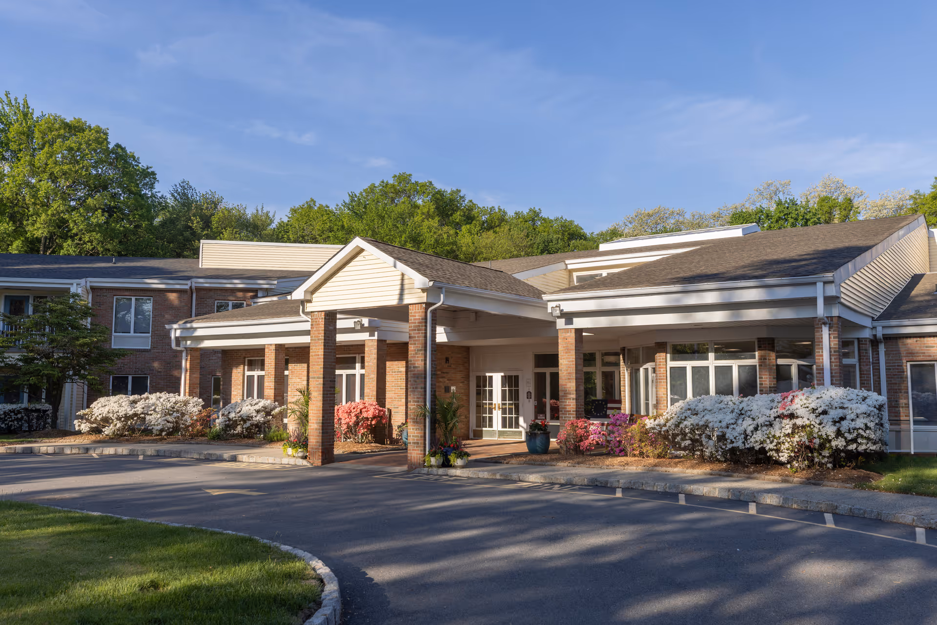 Exterior view of Mira Vie at Montville assisted living facility showing the entrance with a covered driveway, brick pillars, and surrounding flowering bushes and trees under a clear blue sky.