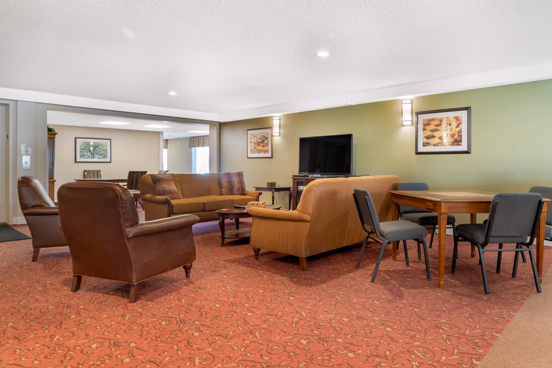 A cozy living room area with two brown leather armchairs, two brown sofas, a wooden coffee table, and a wooden table with four chairs. The walls are painted green and beige, decorated with framed pictures. A flat-screen TV is mounted on a wooden stand against the green wall. The floor is covered with a patterned red carpet.