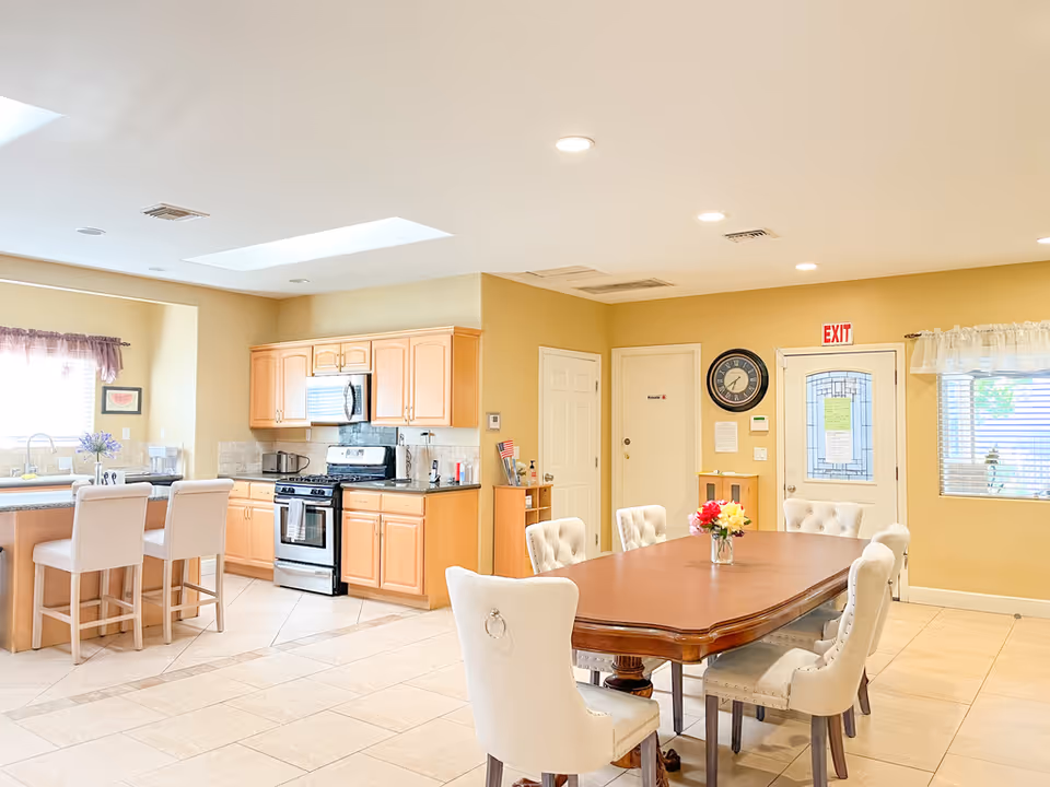 Bright and spacious kitchen and dining area with light wood cabinets, stainless steel appliances, a kitchen island with two white cushioned bar stools, and a wooden dining table surrounded by eight white upholstered chairs. The room has beige tiled floors, recessed ceiling lights, a wall clock, an exit door, and windows with blinds and curtains.