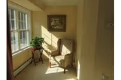 A sunlit corner of a room featuring a patterned armchair next to a small table with a potted plant, with a framed picture hanging on the wall above the chair and large windows letting in natural light.