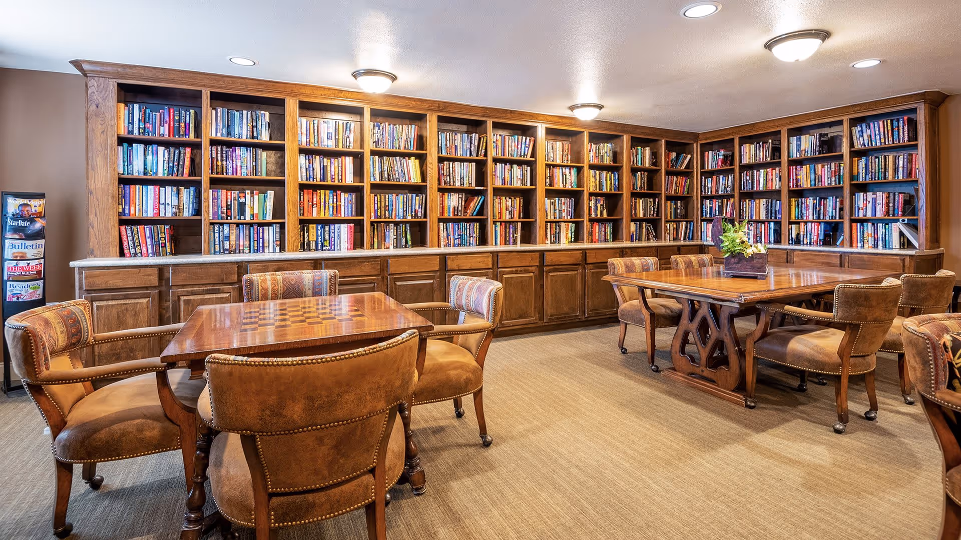 A cozy library room with wooden bookshelves filled with books lining two walls. There are two wooden tables with cushioned chairs around them, one table has a chessboard pattern on its surface. A small flower arrangement is placed on one of the tables. The room has warm lighting and a carpeted floor.