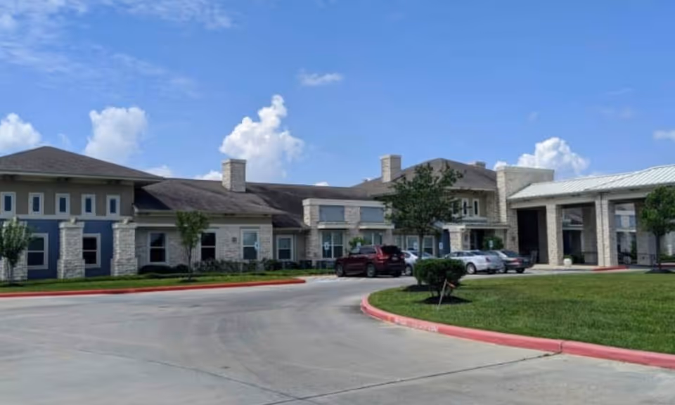 Exterior view of Orchard Park at Victory Lakes Assisted & Senior Living facility showing a single-story building with stone and beige walls, several windows, a covered entrance, a few parked cars, green lawn, and a partly cloudy blue sky.