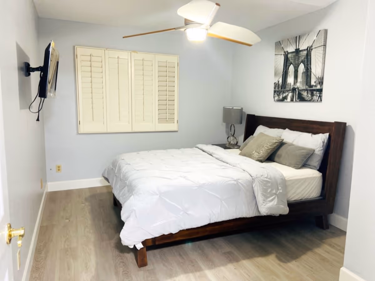 A simple bedroom with a wooden bed made with white bedding and pillows, a nightstand and lamp, ceiling fan, wall-mounted TV, window shutters, and framed wall art.
