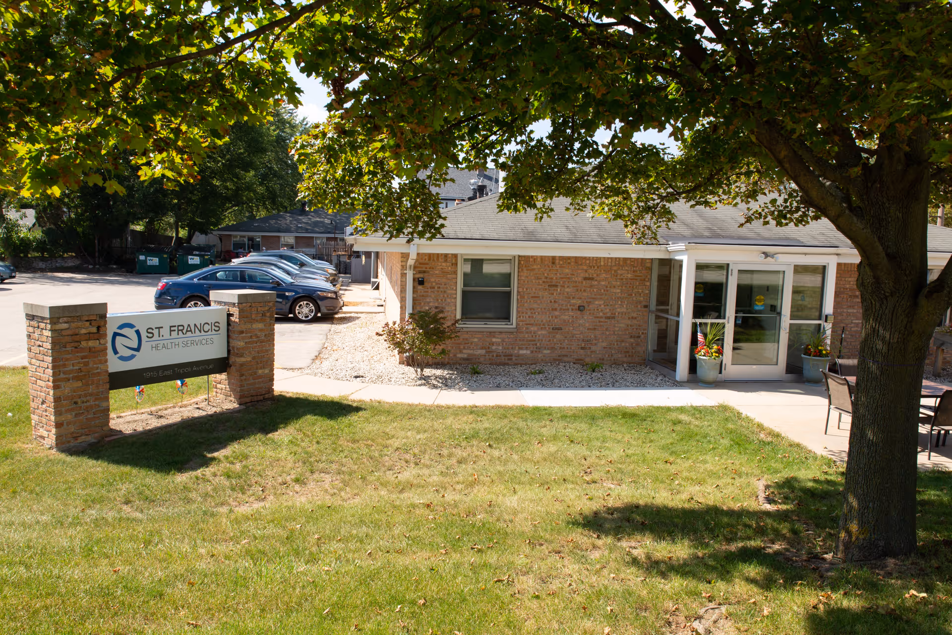Exterior view of Saint Francis Health Services building with a brick facade, a glass entrance door, a green lawn, a large tree providing shade, and a parking lot with several cars in the background.