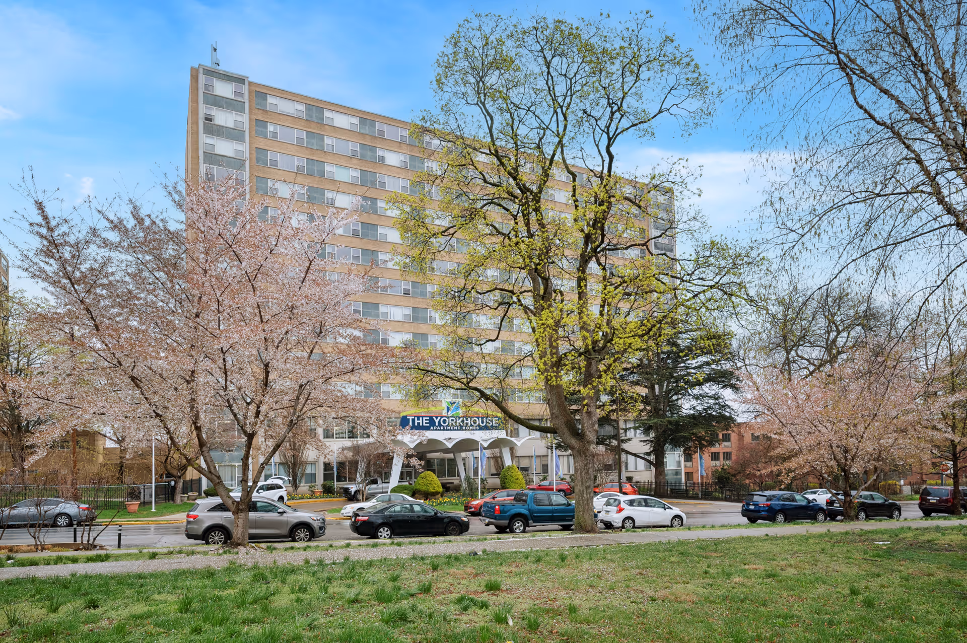 Exterior view of a multi-story apartment building named The York House Apartments, surrounded by trees with spring blossoms and parked cars along the street in front.