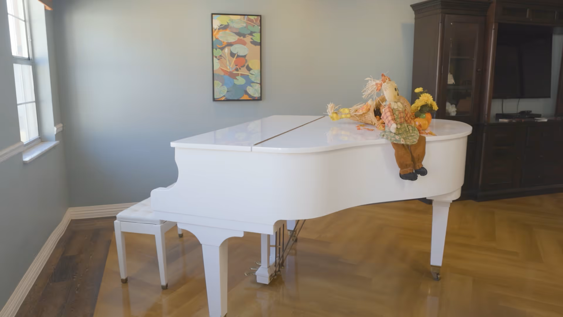 A bright room with a white grand piano and matching white piano bench on a wooden floor. On top of the piano, there is a decorative scarecrow doll and some yellow flowers. A colorful framed artwork hangs on the light blue wall, and a dark wooden cabinet with a TV is visible in the background.