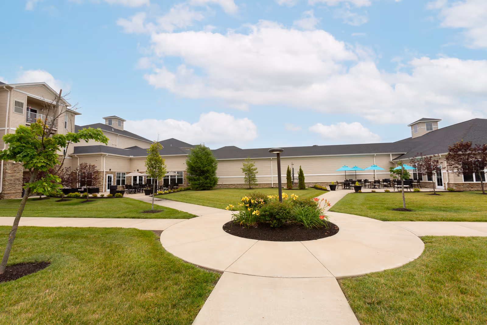 Outdoor courtyard area of a senior living facility with a circular concrete walkway surrounding a flower bed, green grass, small trees, and patio tables with blue umbrellas. The building has beige siding and multiple windows under a partly cloudy sky.