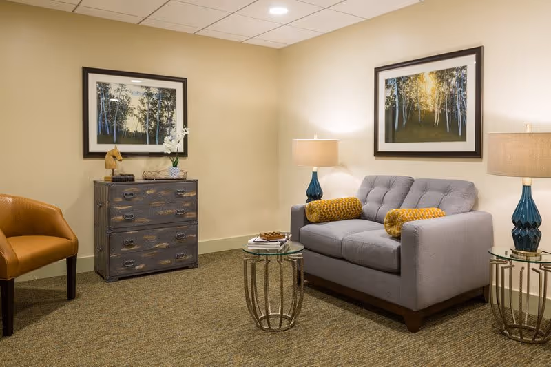 A cozy living room area with a gray tufted sofa adorned with two yellow patterned bolster pillows, flanked by two glass-top side tables each holding a blue lamp with a beige shade. To the left, there is a brown leather armchair and a rustic wooden chest of drawers with decorative items on top. Two framed pictures of trees hang on the beige walls.
