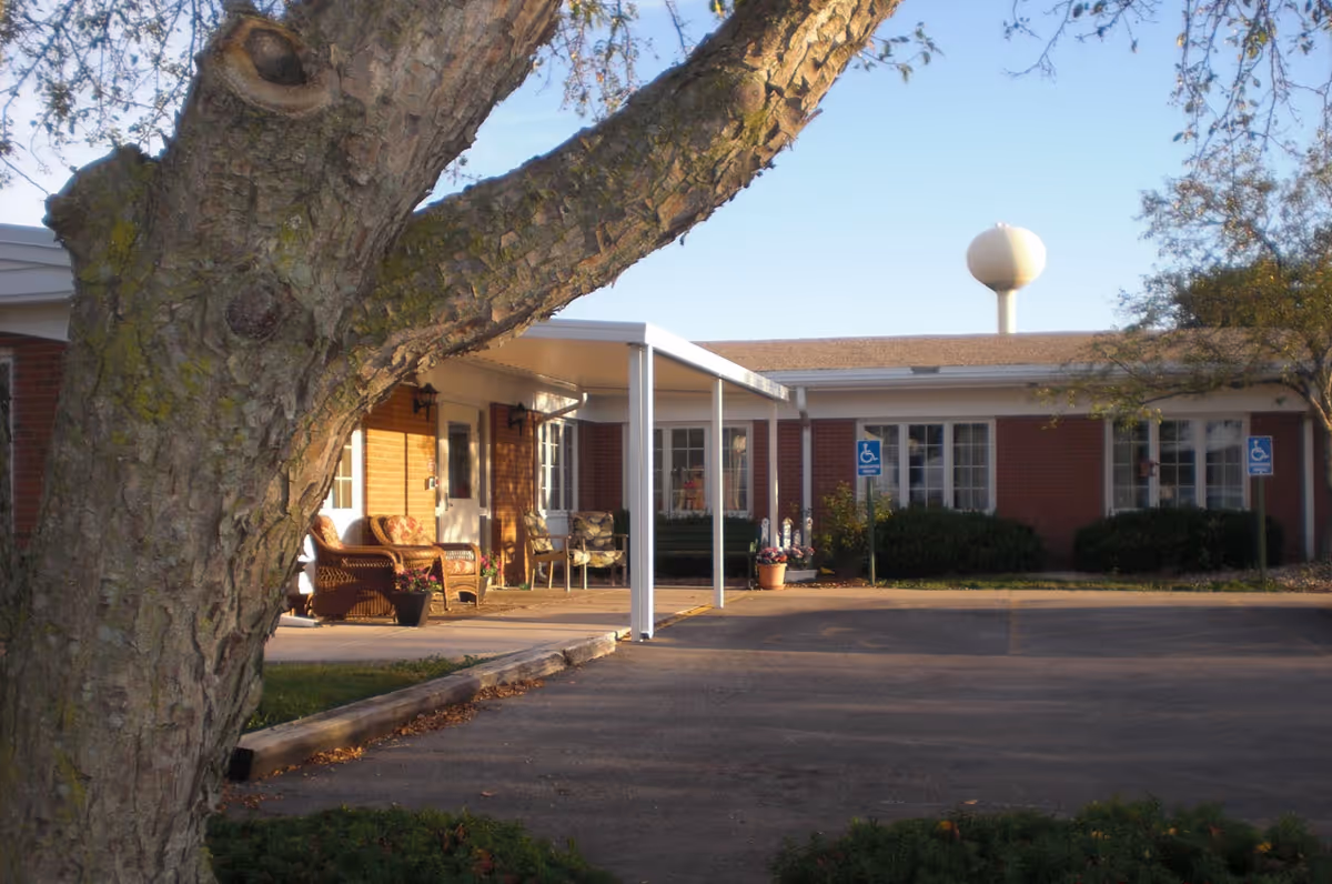 View of the front exterior of a single-story brick building with a covered entrance and outdoor seating area. There is a large tree in the foreground and two handicap parking signs near the entrance. A water tower is visible in the background under a clear sky.