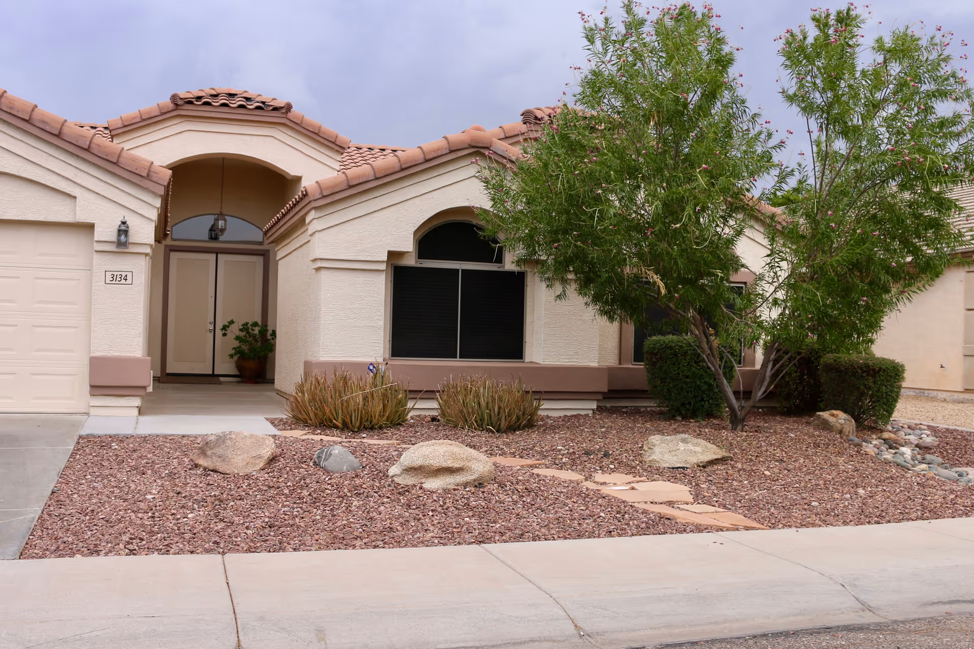 Exterior view of a single-story residential building with beige stucco walls and a tiled roof. The entrance features a covered porch with double doors and a hanging light fixture. The front yard is landscaped with rocks, a few bushes, a tree, and a stone pathway leading to the entrance. The house number 3134 is visible next to the garage door.