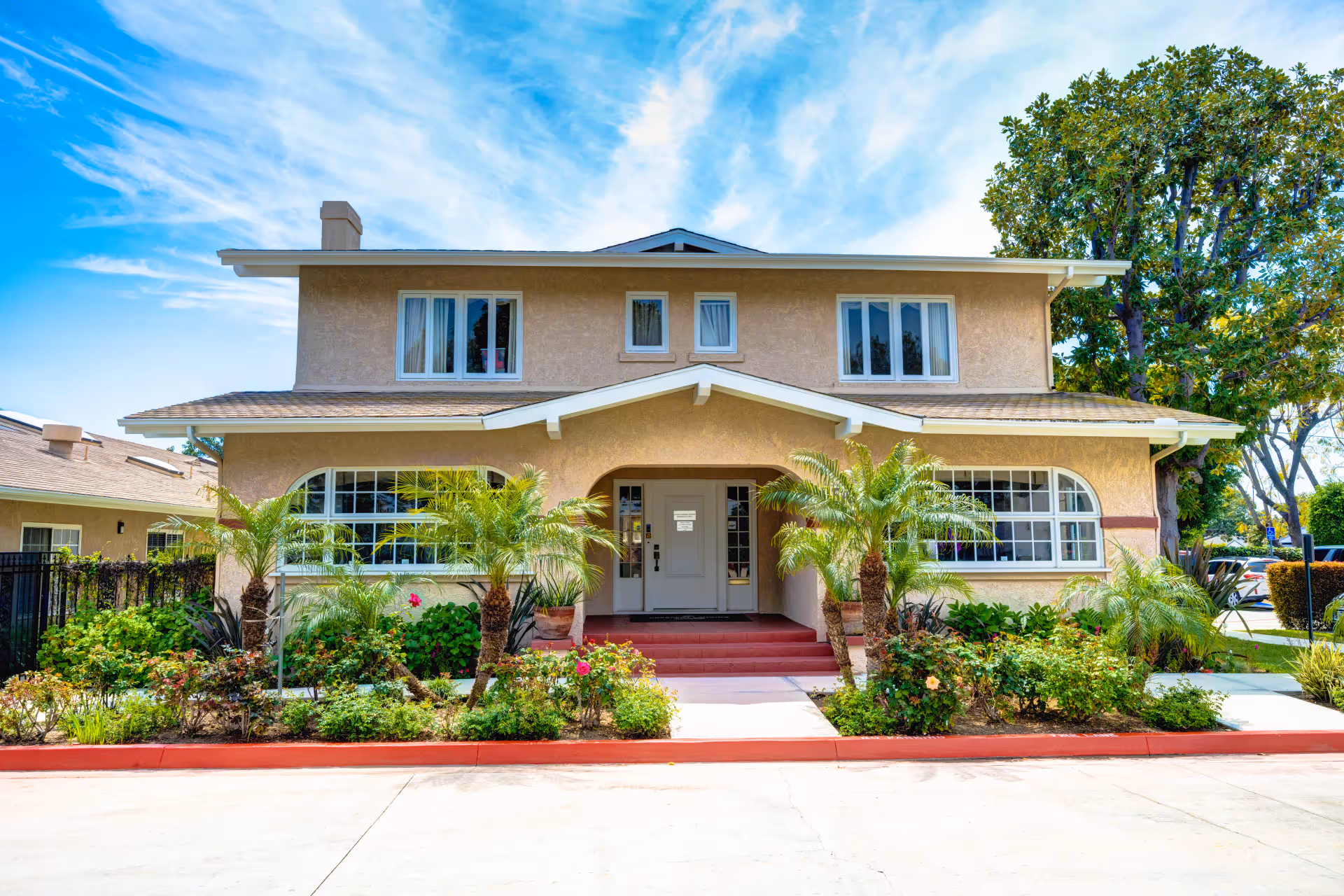 Front view of a two-story beige stucco building with a central entrance, palm trees, and landscaped flower beds.