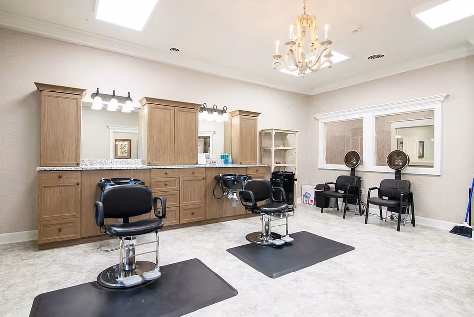 Interior of a hair salon area with two black salon chairs on black mats in front of wooden cabinets with sinks and mirrors. Two black hair dryer chairs are positioned against the wall with large windows above them. The room has light-colored walls and flooring, with a chandelier hanging from the ceiling.
