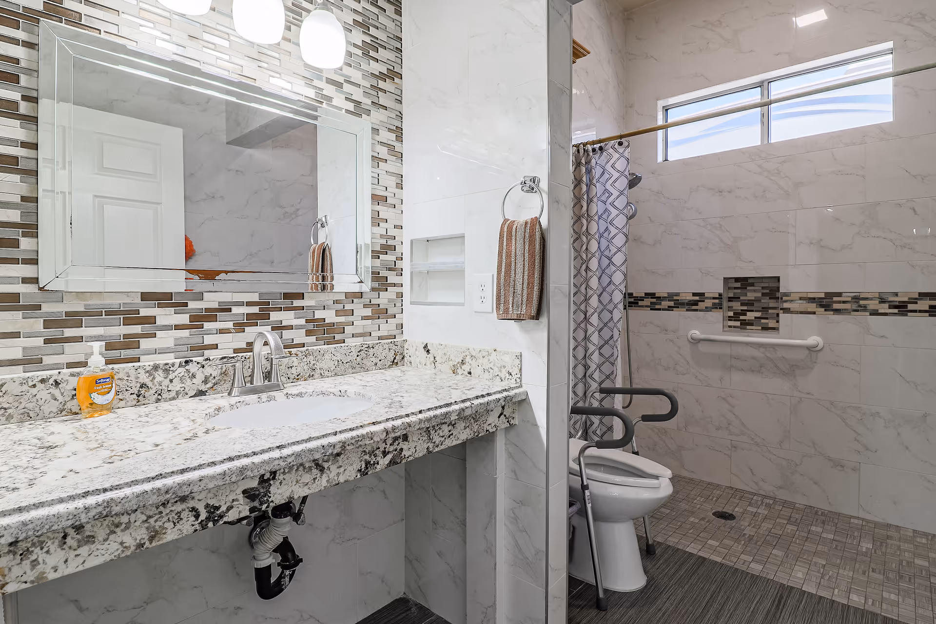 Accessible bathroom featuring a granite countertop with sink and mirror, and a roll-in shower with grab bars and shower chair.