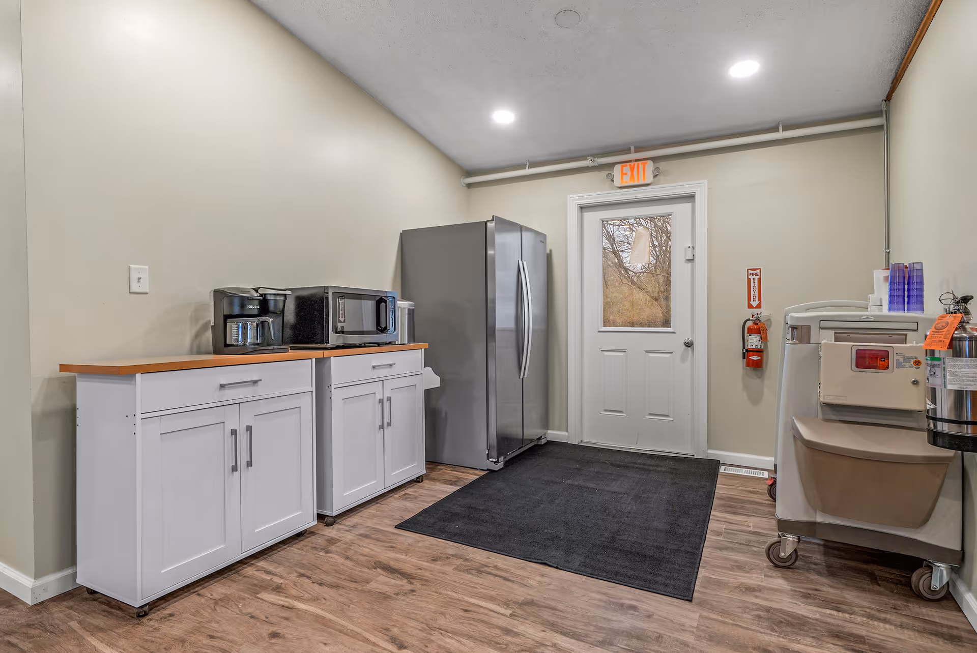 A small kitchen area with white cabinets, a coffee maker, microwave, and a large stainless steel refrigerator. There is a door with a window leading outside, an exit sign above the door, a fire extinguisher mounted on the wall, and a large black mat on the wooden floor. The walls are painted beige and the ceiling has recessed lighting.