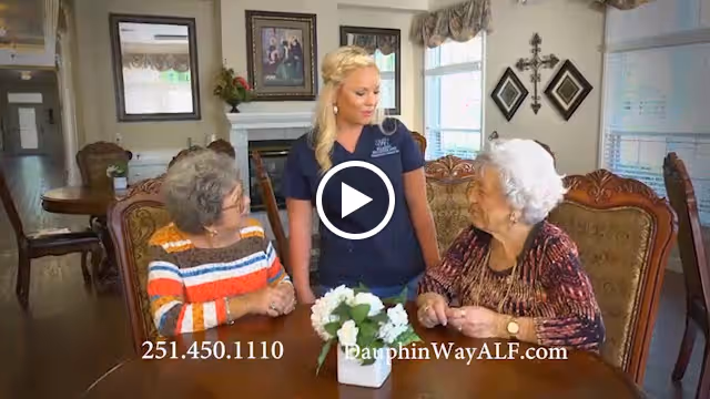 A caregiver speaks with two elderly women seated at a dining table in a decorated senior living common area.