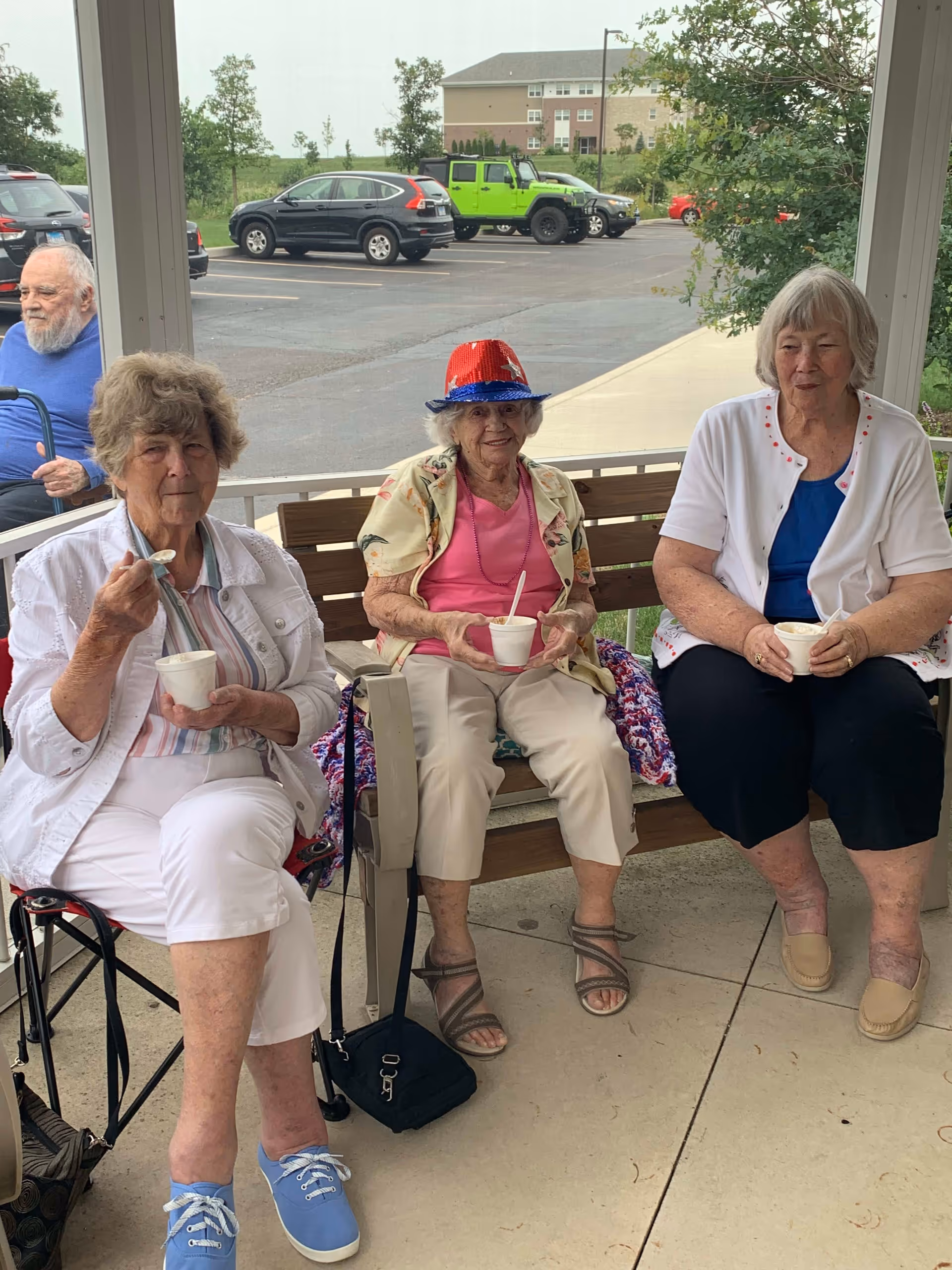 Three elderly women sitting outside on a covered patio, each holding a cup with a spoon. One woman in the middle is wearing a red, white, and blue hat. There is a man in a wheelchair in the background and several parked cars in a parking lot behind them.