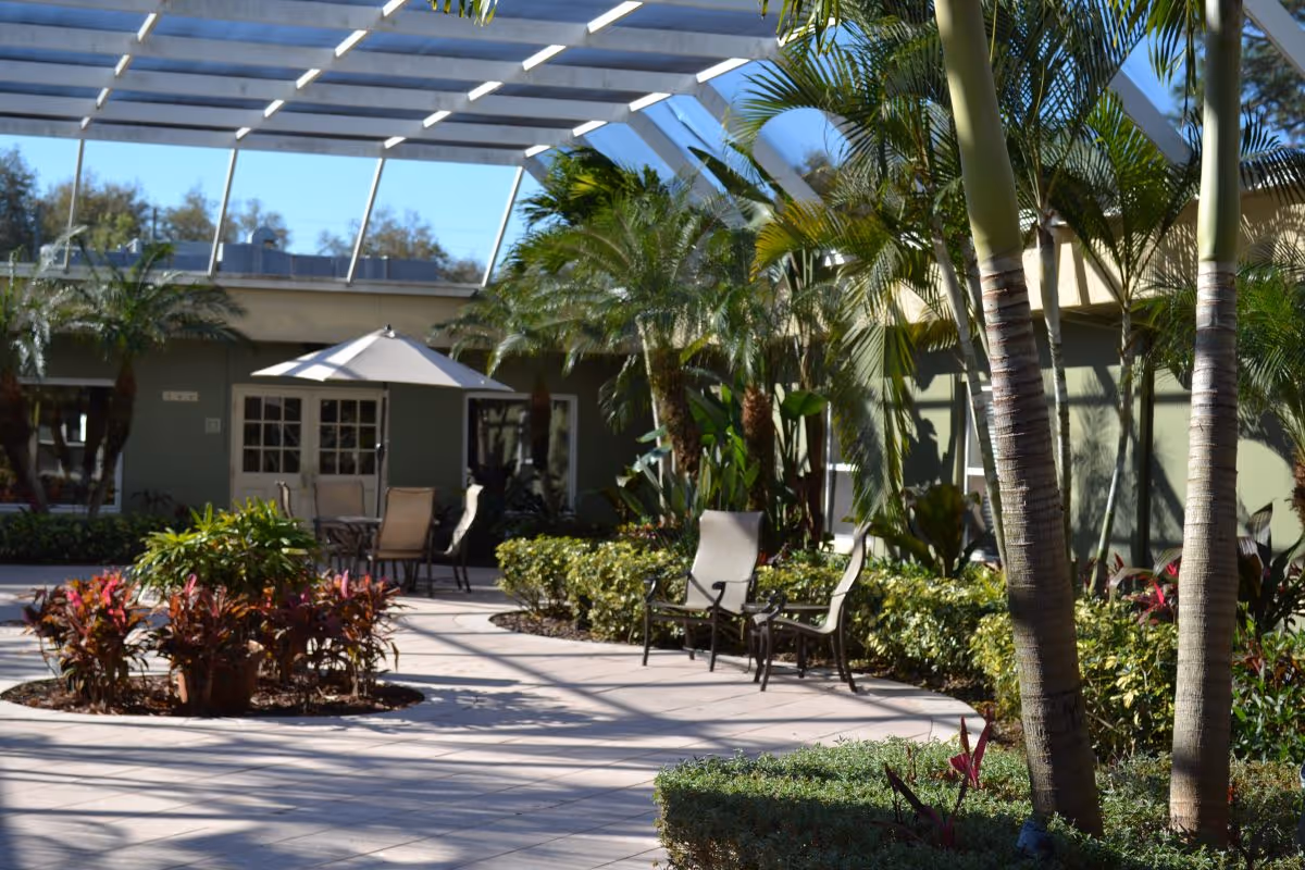 Sunlit enclosed courtyard with patio tables, chairs and an umbrella surrounded by palm trees and landscaping.