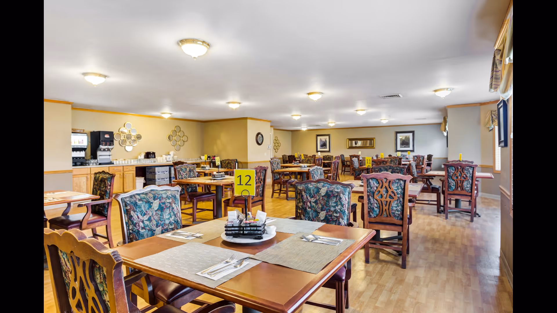 A spacious dining room with multiple wooden tables and chairs arranged neatly. Each table has placemats, cutlery, and a small centerpiece. The room has light-colored walls with decorative mirrors and framed pictures. There is a beverage station with a coffee machine and cups along one wall. The floor is wooden, and the ceiling has multiple light fixtures providing bright illumination.