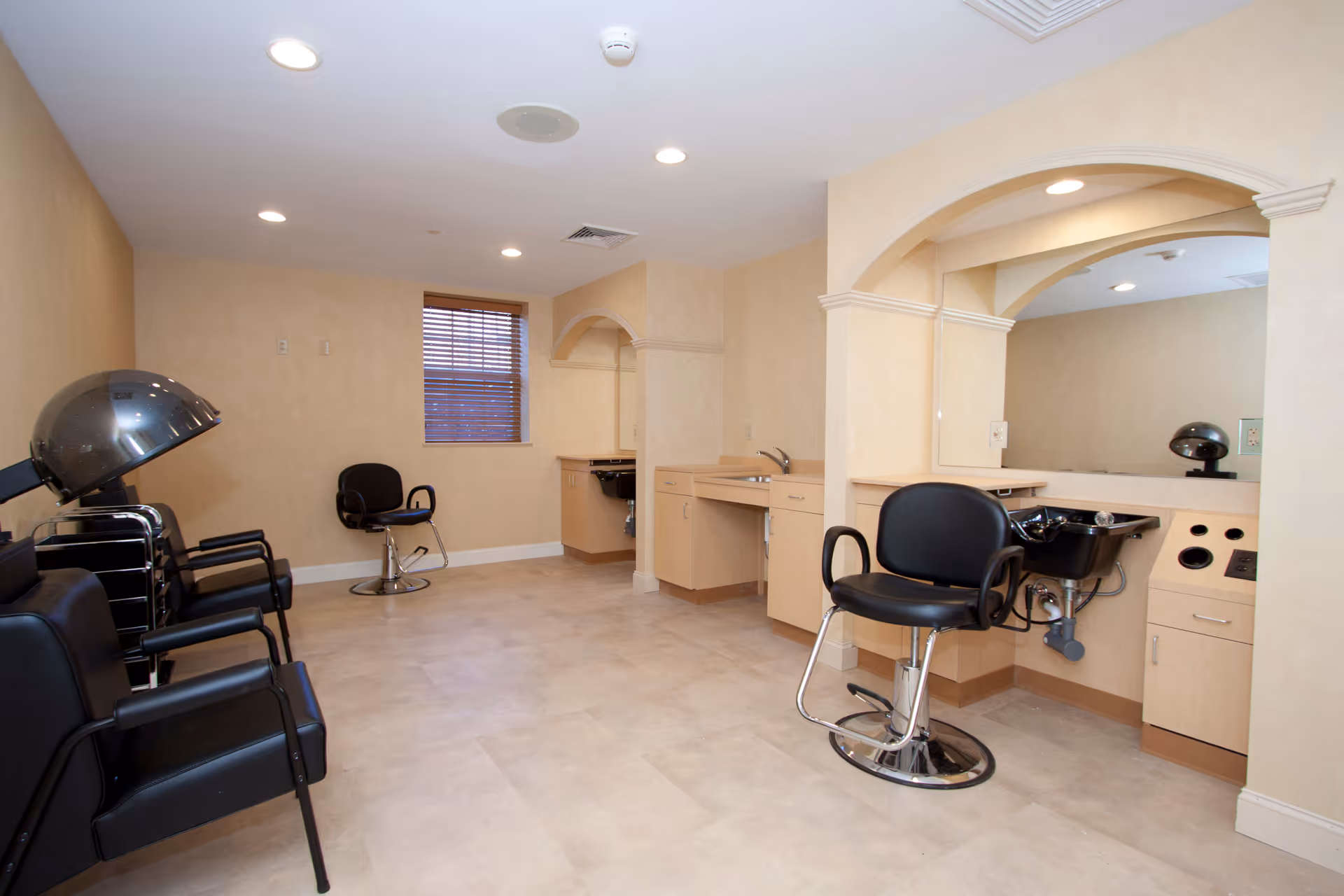 A salon room with styling chairs, hooded hair dryers and shampoo sinks in a neutral-toned interior.