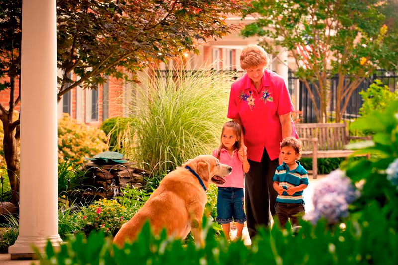 An elderly woman in a bright pink shirt stands outdoors with two young children, a girl and a boy, near a large golden retriever dog in a garden area with green plants, flowers, and a bench in the background.