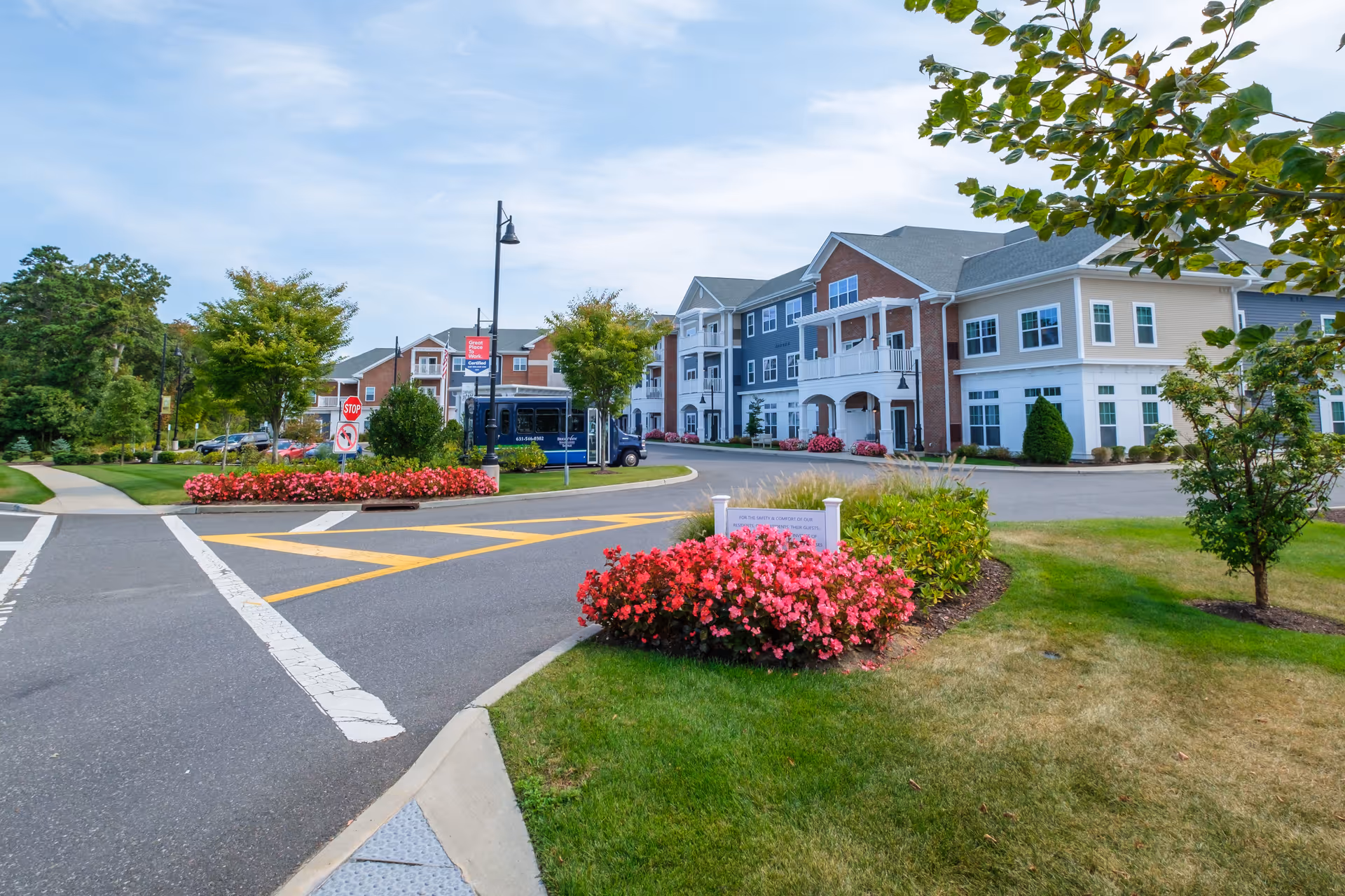 Exterior view of a senior living facility with a multi-story building featuring balconies, surrounded by well-maintained landscaping including green lawns, trees, and pink flowering bushes. A paved driveway with road markings and a stop sign is visible, along with a blue shuttle bus parked near the building.