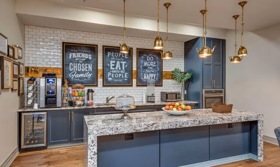 A modern kitchen area with a large granite island countertop in the foreground. The back wall features dark blue cabinets, a coffee machine, a small refrigerator, a microwave, and a water dispenser. Three framed chalkboard-style signs with inspirational food and friendship quotes hang on a white subway tile backsplash. Five brass pendant lights shaped like teapots hang from the ceiling above the island. A bowl of assorted fruit and a basket are placed on the island countertop.