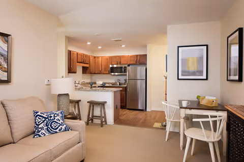 Open-plan living area with a sofa, small dining table, and a kitchen with wooden cabinets and stainless-steel appliances.