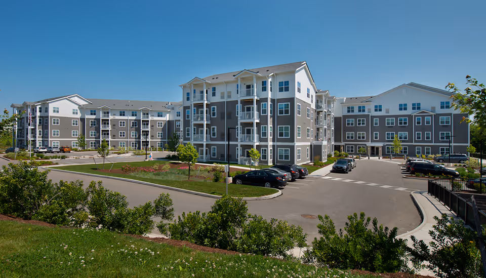 Exterior view of The Province of Wexford senior living facility showing a large, modern multi-story building with gray and white siding, multiple balconies, a parking lot with cars, landscaped greenery, and a clear blue sky.
