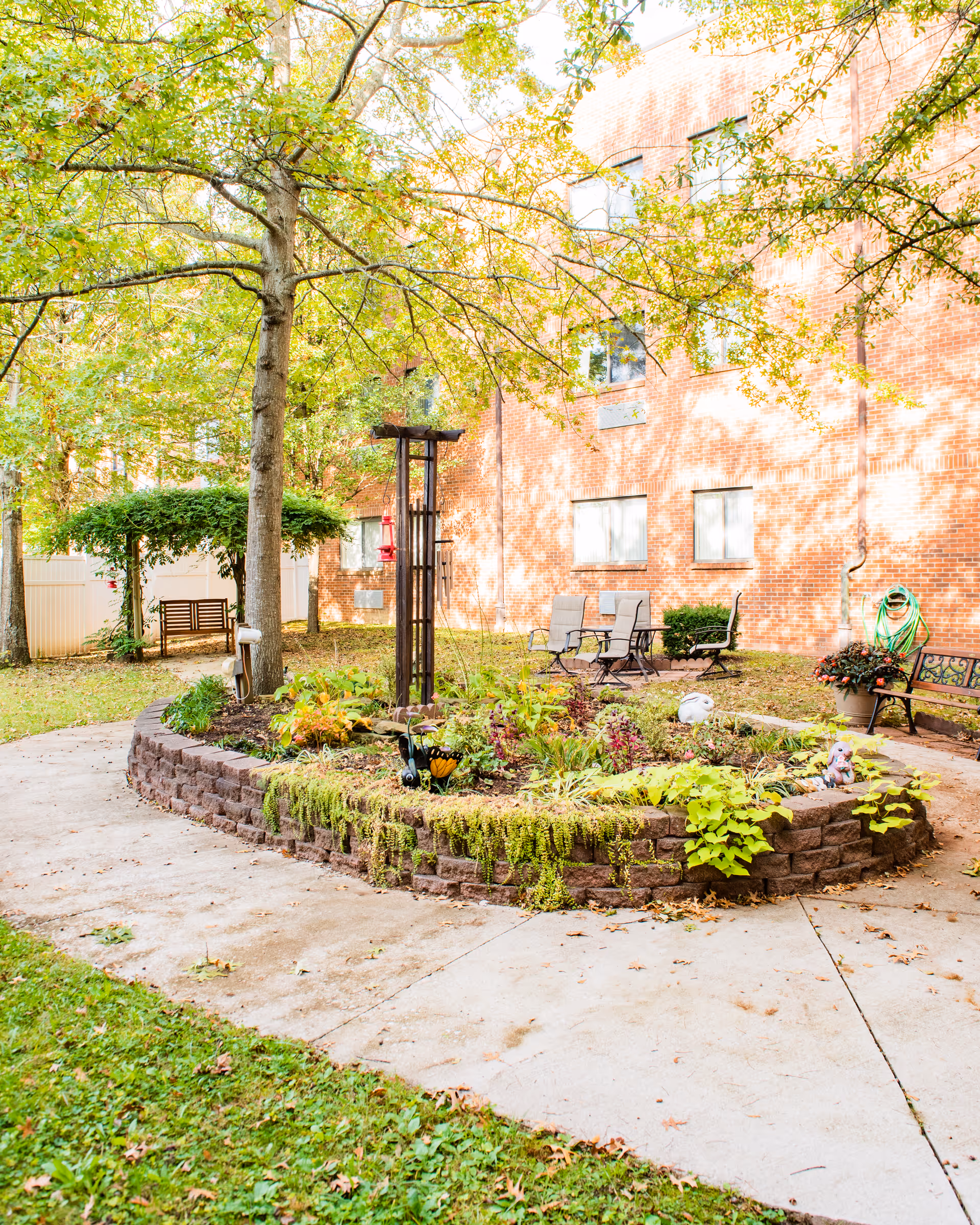 A sunny courtyard with a circular raised flower bed, patio chairs and benches in front of a brick building.