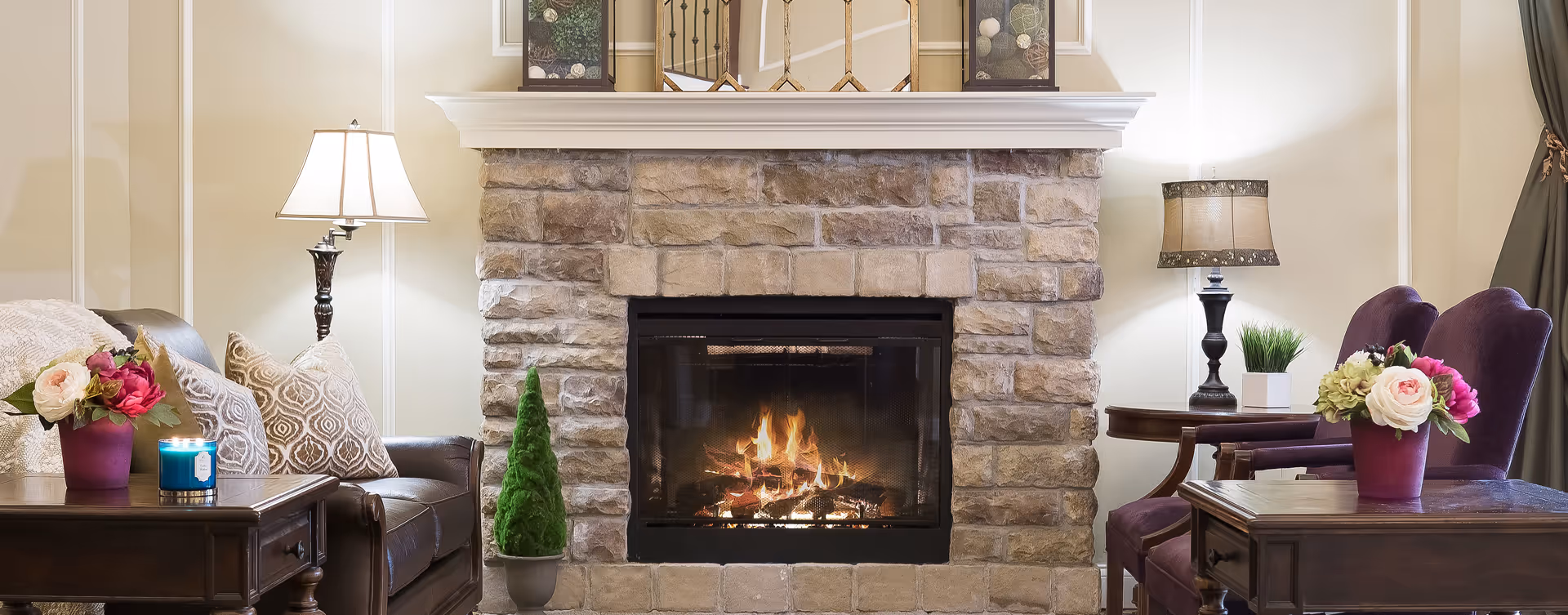 Cozy living room with a stone fireplace flanked by armchairs, side tables, lamps, and floral arrangements.