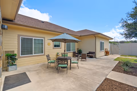Outdoor patio area at Poet's Walk San Antonio, A Memory Care Community, featuring a round table with four chairs and a large umbrella. The patio is adjacent to a beige building with windows and a brown roof. There are some plants in pots and a small seating area with chairs along the building wall. The sky is clear with a few clouds.