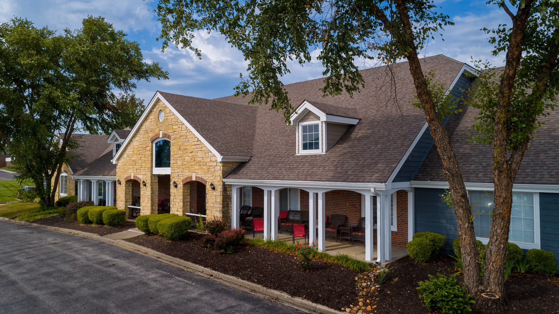 Front façade and covered porch of a single-story assisted living building with stone accents, chairs on the porch, trees, and landscaped beds.