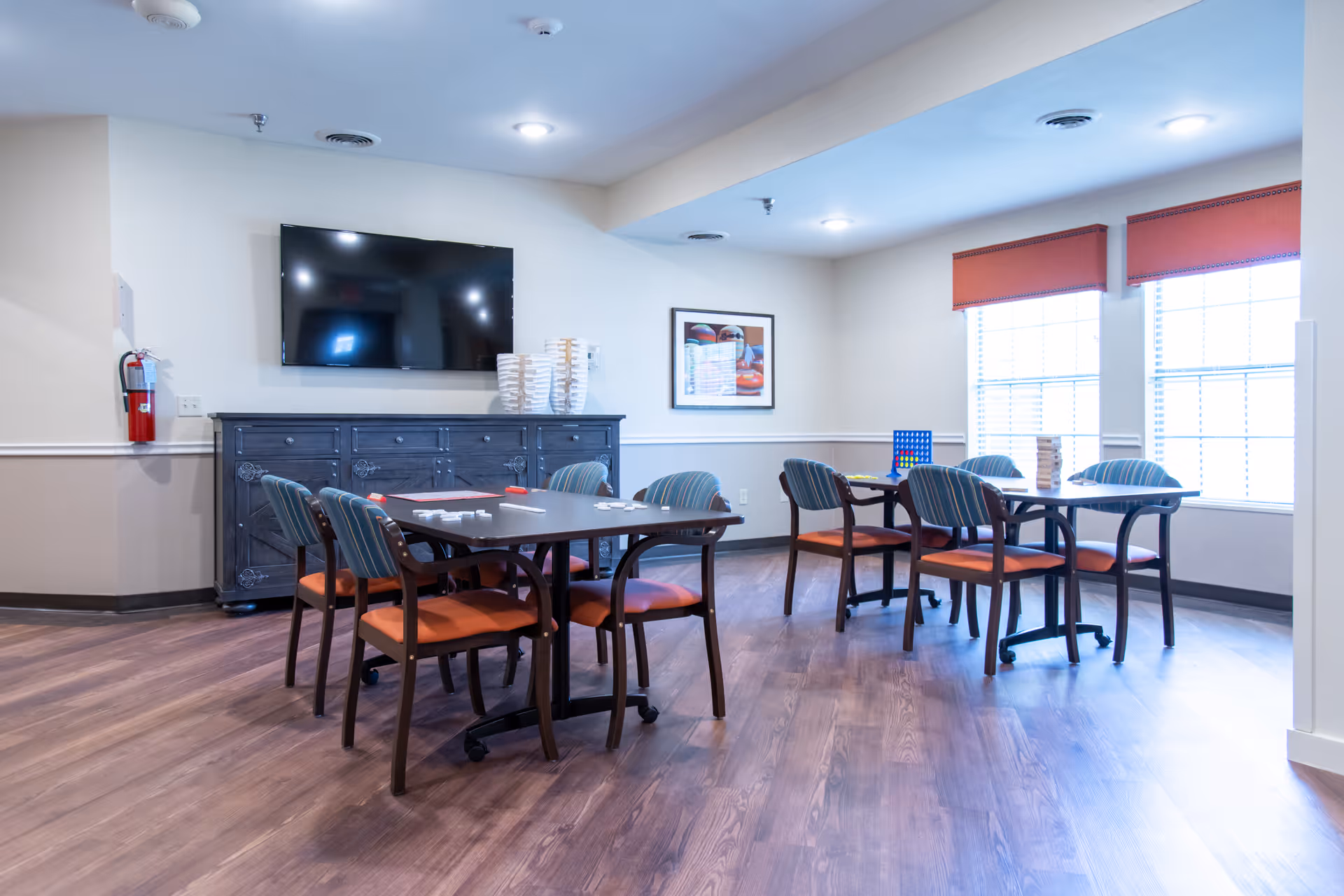 A bright room with two tables and eight chairs arranged around them. The chairs have striped backs and orange seats. On the tables are board games including Jenga and Connect Four. A large flat-screen TV is mounted on the wall above a dark wooden cabinet. Two windows with red valances let in natural light. The floor is wood and the walls are painted light beige with a white chair rail.